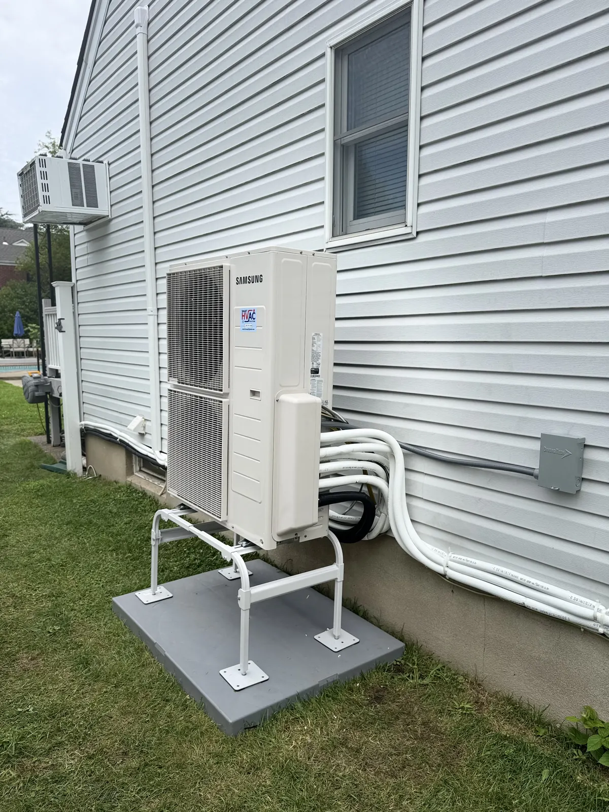 Outdoor mini-split condenser on a raised stand beside a home with multiple refrigerant line sets running to the wall